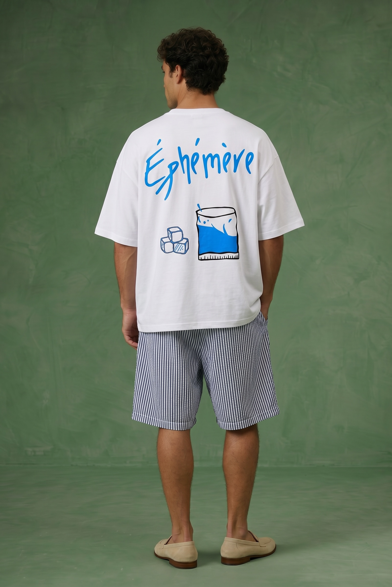Person wearing a white t-shirt with blue text and graphics on the back ephemere standing against a green background.